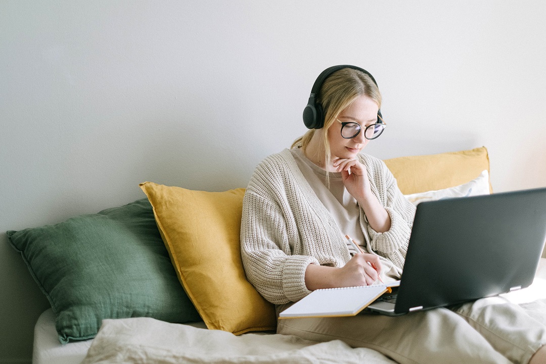 Mujer con audífonos trabajando con libreta y laptop en cama