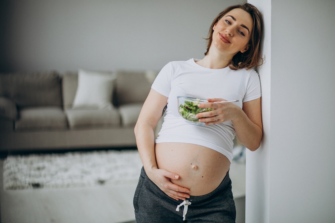 Este artículo tiene un ejemplo práctico de un día de comida que te puede hacer más fértil, incluyendo alimentos si eres vegetariana o vegana. *** Mujer embarazada comiendo ensalada en cocina