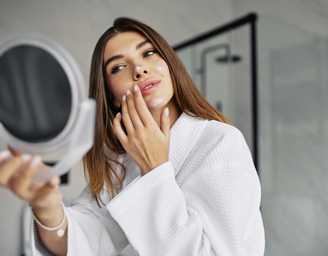 Mujer cuidando su cara skincare crema rutina belleza espejo bata