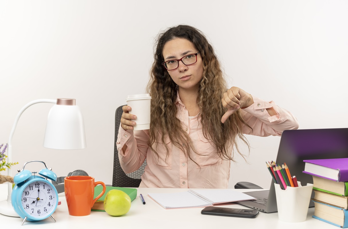 Joven disgustada con gafas escritorio tarea trabajo taza café pulgar hacia abajo