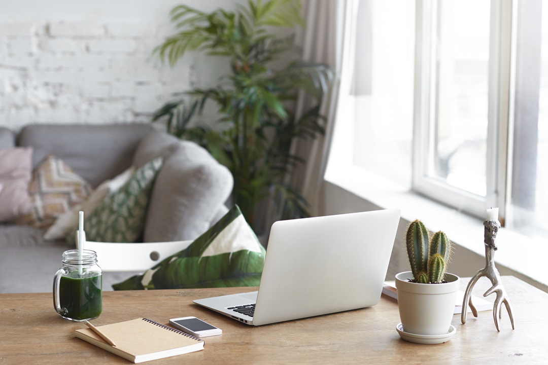 Workplace of modern freelancer leading healthy lifestyle: open generic laptop computer, cactus in pot, smart phone, copybook and glass of green smoothie on wooden desk in stylish home office interior